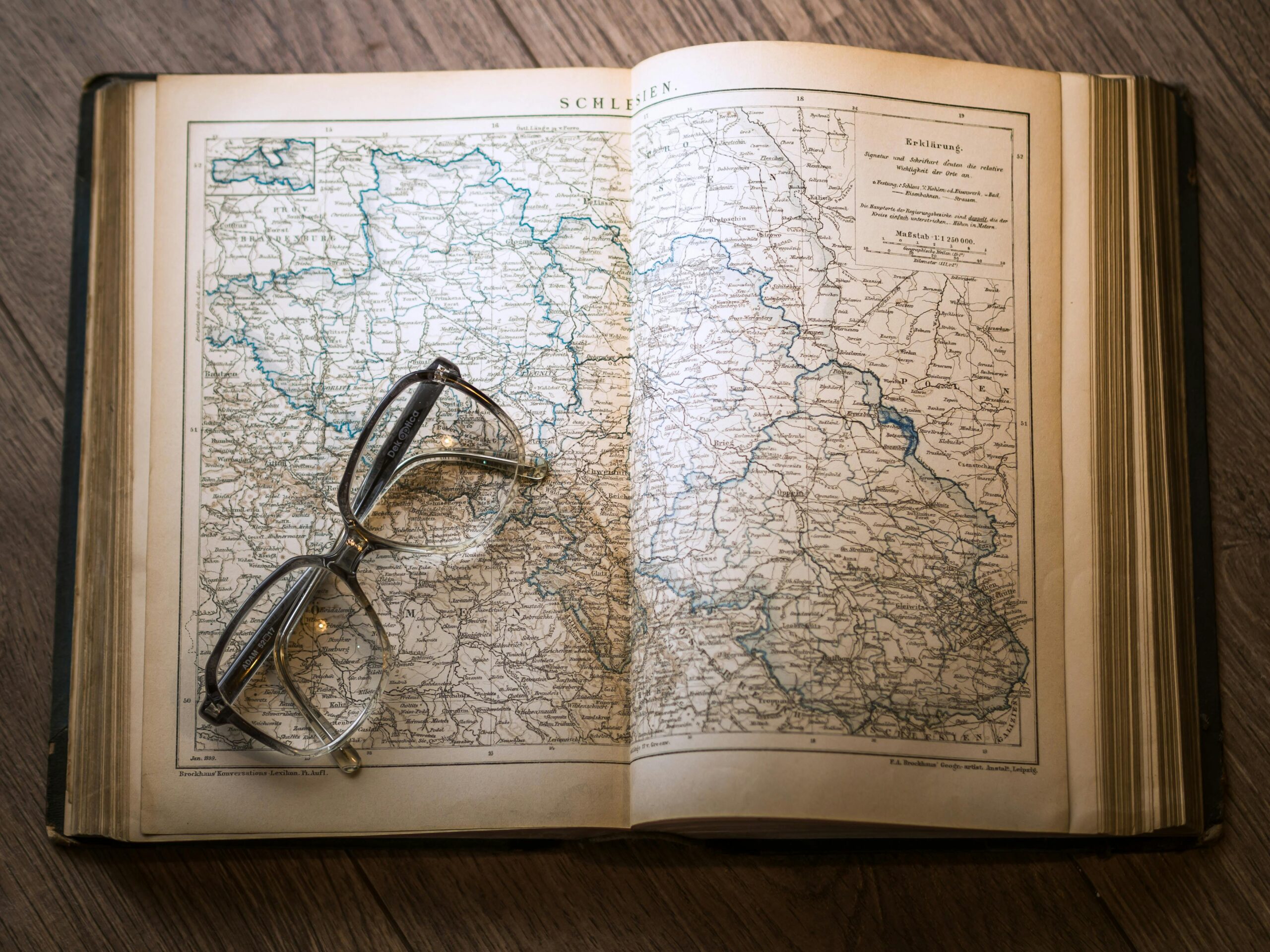 A close-up of a vintage map with eyeglasses resting on an open book, set on a wooden table.